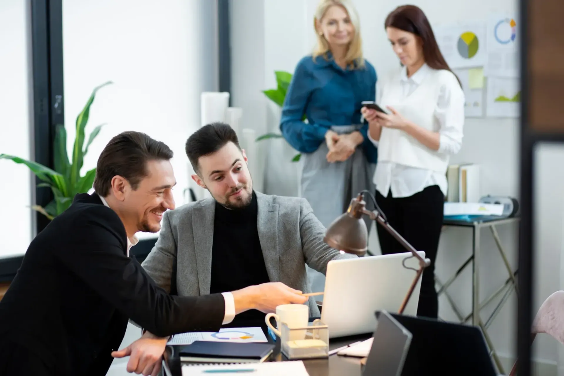 Business colleagues collaborating at a laptop in office; two men point at screen, two women look on.