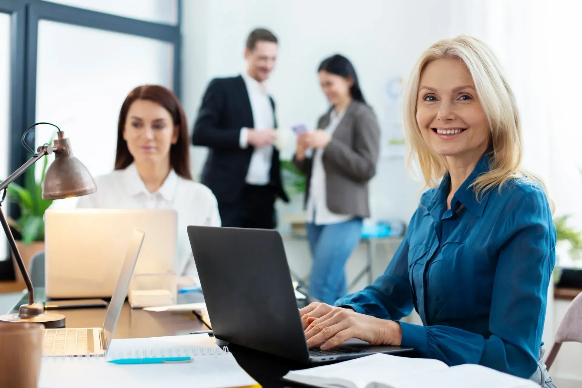 Woman smiling at camera while working on a laptop at a desk, with coworkers in the background.