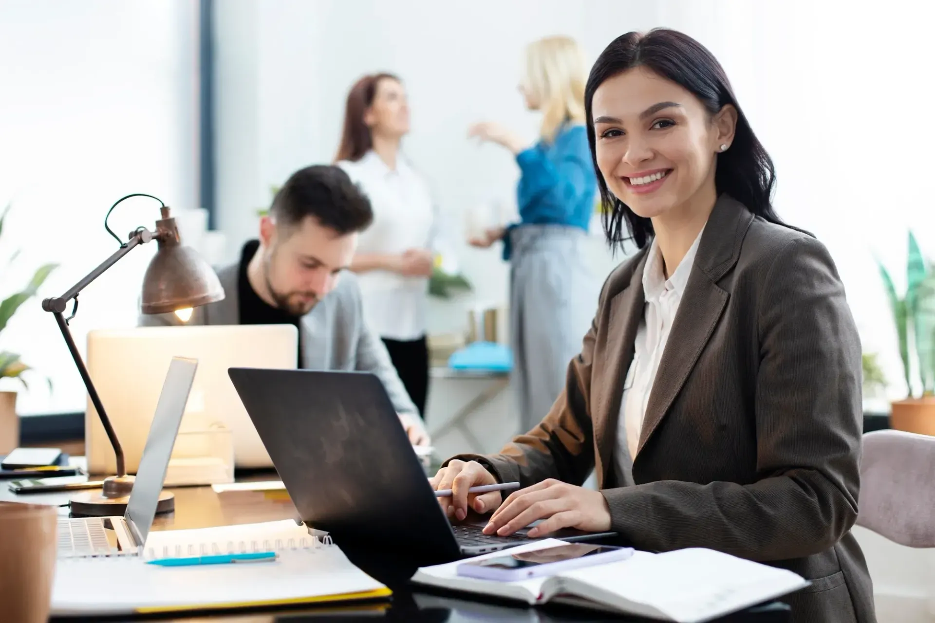 Woman in blazer working on laptop, smiling in office.