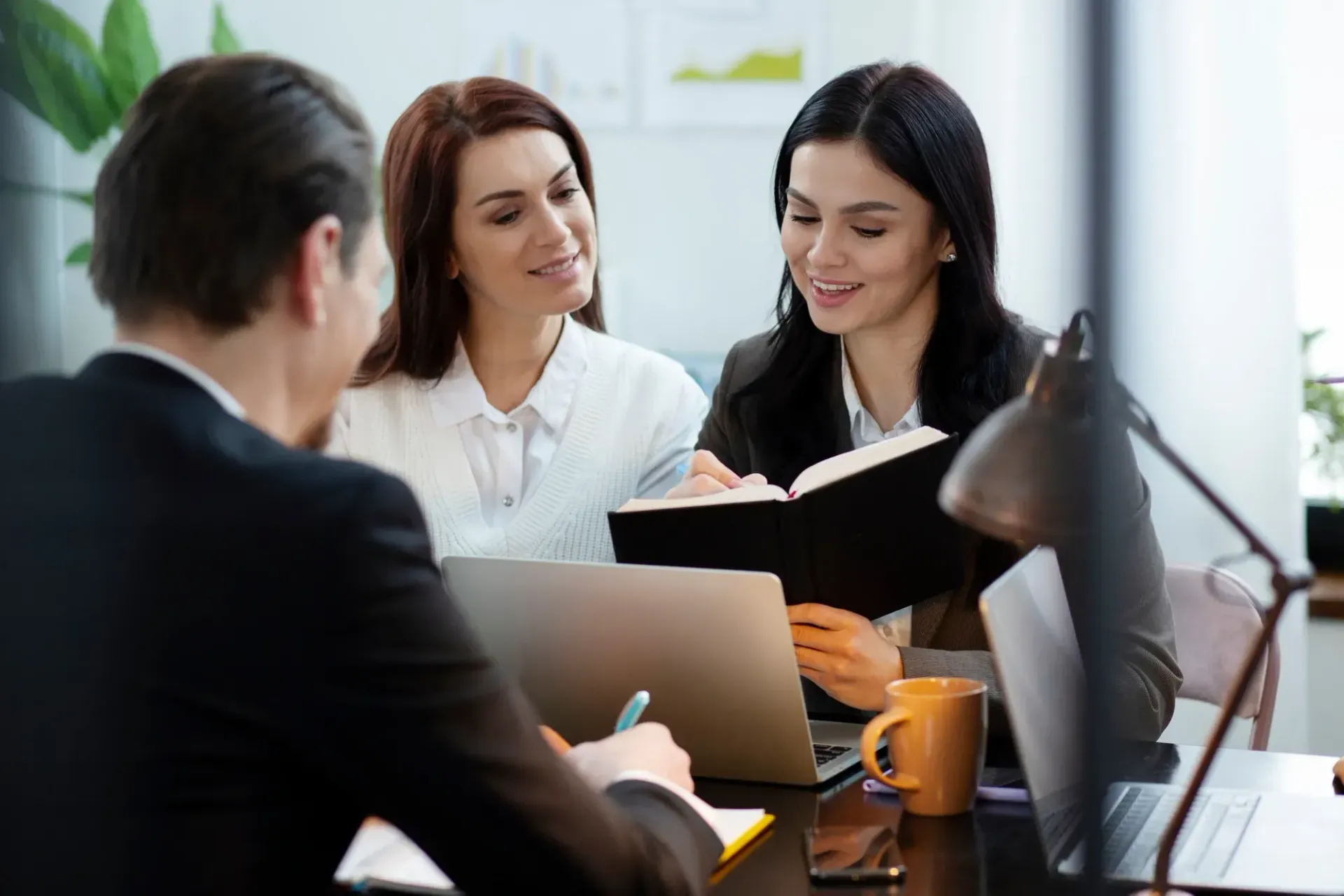Three people in business attire at a desk, reviewing documents and a laptop, lit by a lamp.