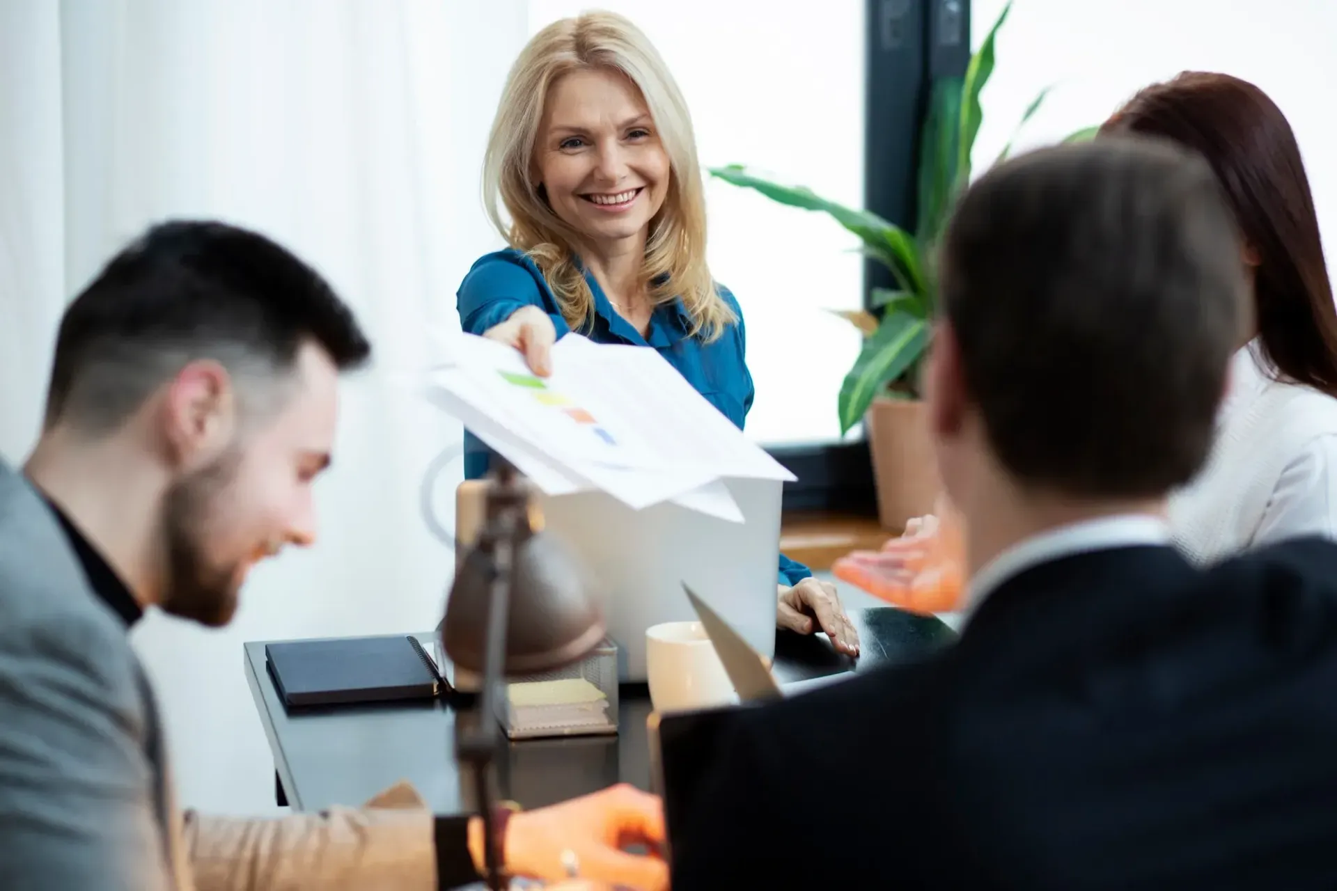Woman in blue shirt handing documents to colleagues at a table; they are smiling.