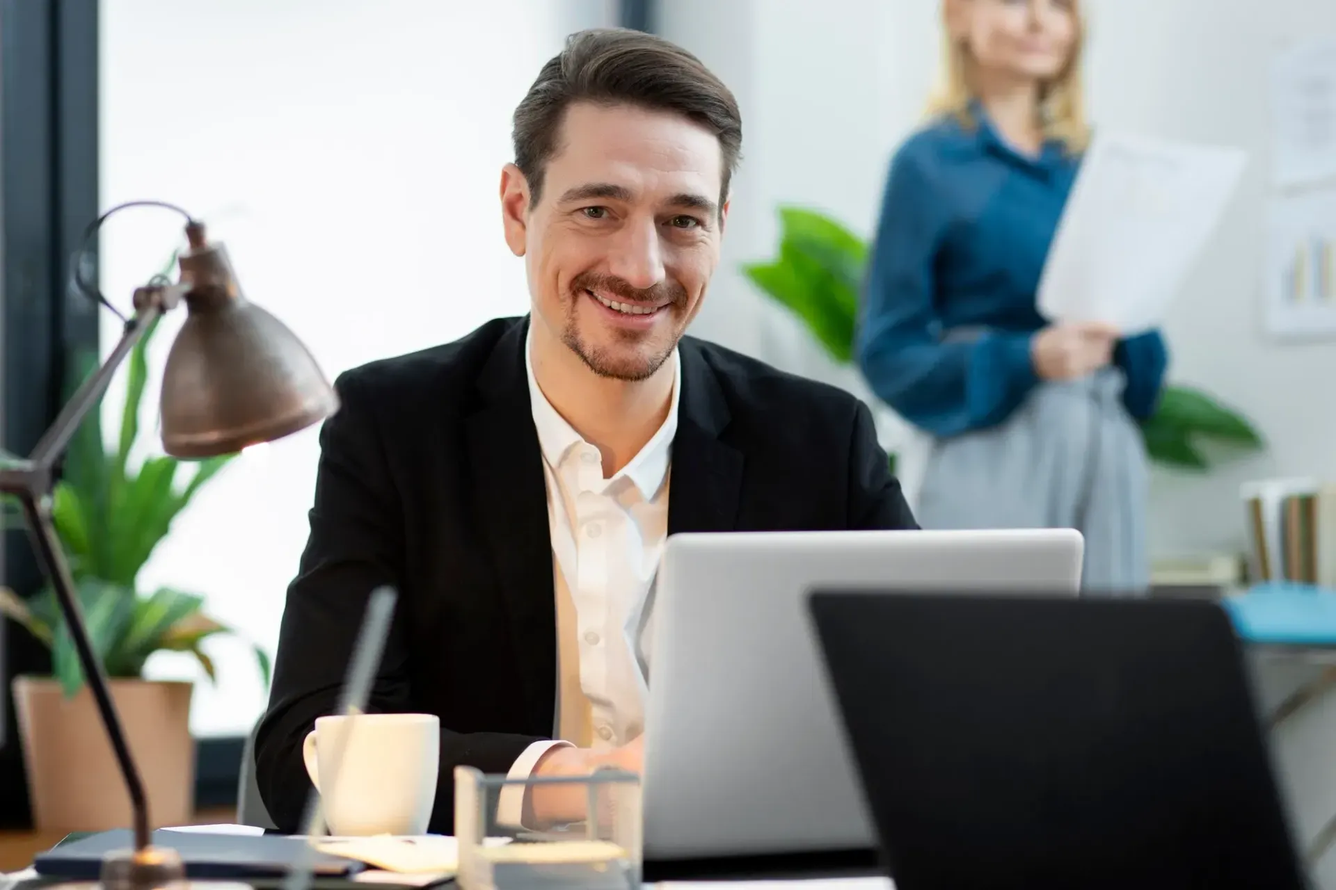 Man in a black suit smiles at a desk, working on a laptop. A woman stands in the background, holding papers.