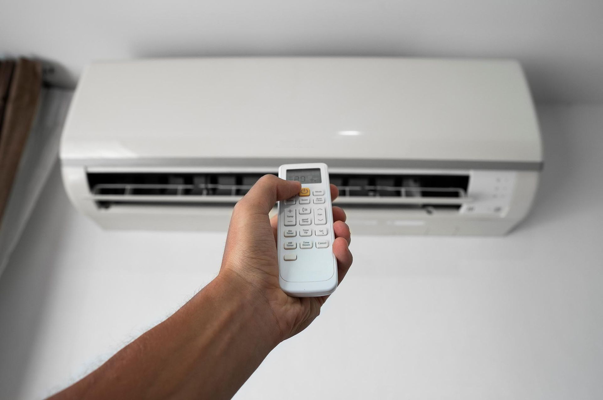 Hand holding a remote pointed at a wall-mounted air conditioner in a white room