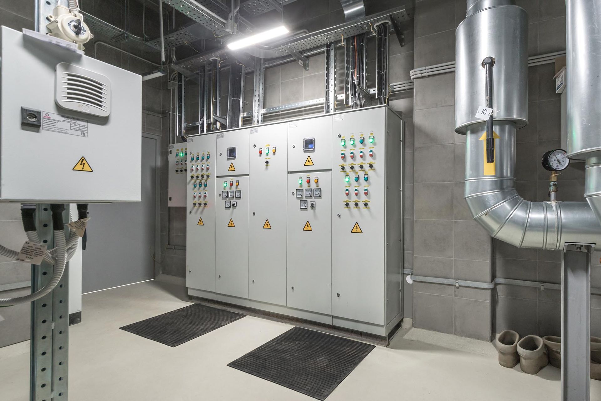 Industrial electrical control room with gray cabinets, warning signs, ducts, and pipes in a utility basement