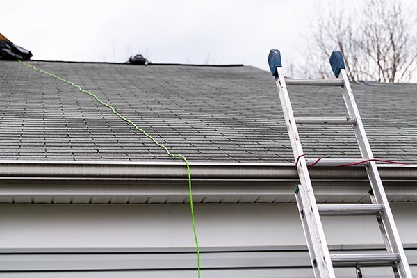 Ladder leaning against a gray shingled roof, green cord running down the side. White siding below. Ladder leaning against a gray shingled roof, green cord running down the side. White siding below.