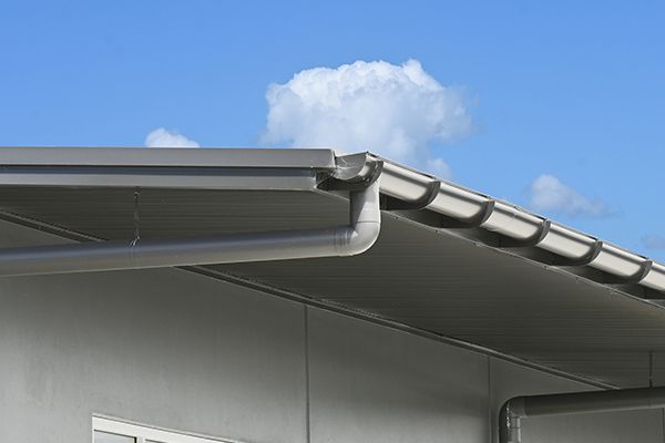 Gray roof edge with gutter and downspout against a blue sky with fluffy clouds. Gray roof edge with gutter and downspout against a blue sky with fluffy clouds.