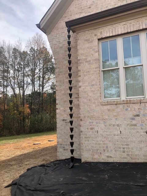 A dark brown rain chain attached to a beige brick house, leading to a black ground covering.