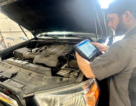 Mechanic using a diagnostic tablet under the open hood of a car in a garage.