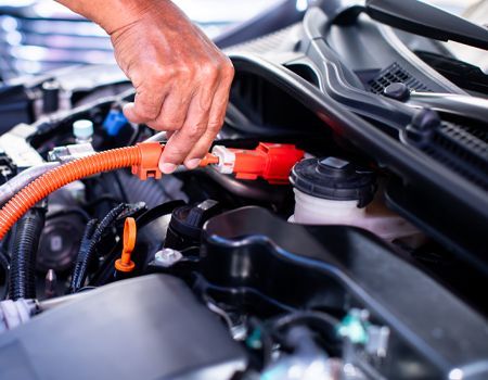Person's hand connecting an orange charging cable to a red positive terminal in a car engine bay.