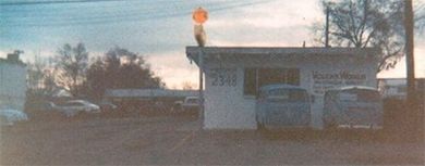 An old gas station building with cars parked in front, topped with a traffic signal. The sky is overcast.