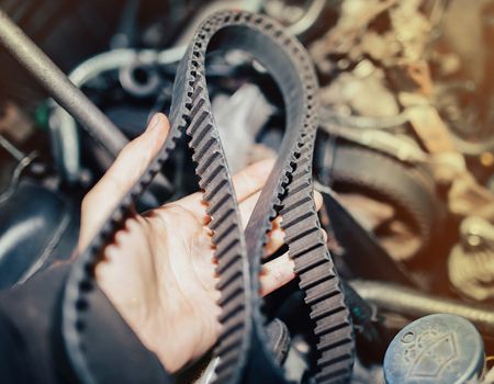 A hand holding a worn, toothed timing belt in front of an engine, likely for inspection or replacement.