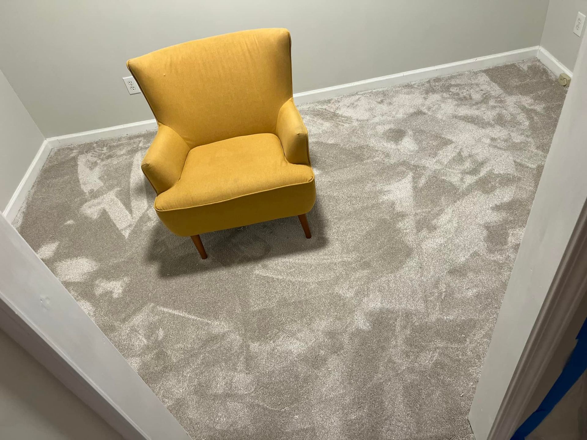 Yellow armchair in a light gray, patterned carpeted room. White trim borders the room, which appears to be a small, angled space.