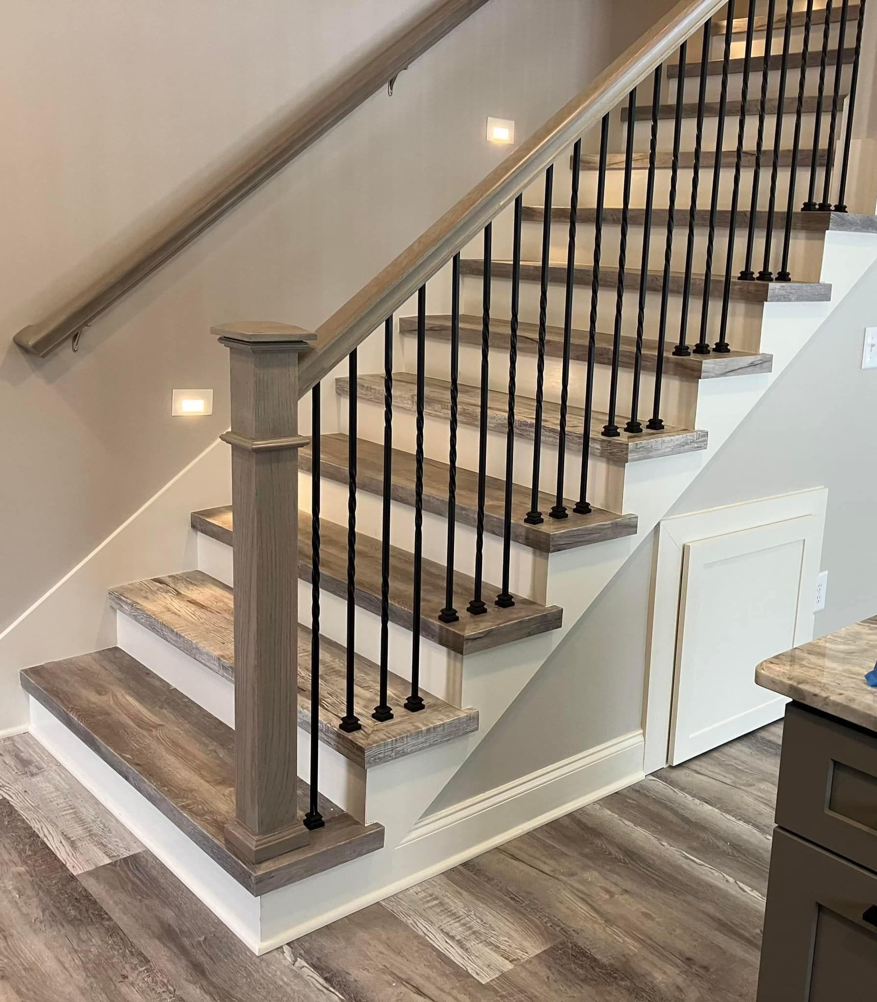 Staircase with gray wood-look steps, white risers, and black iron spindles. Beige walls and a matching handrail.