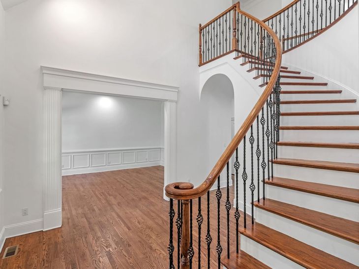 A grand wooden staircase with a curved railing leads to an upper level.  A doorway opens into a room with wood floors and white walls.