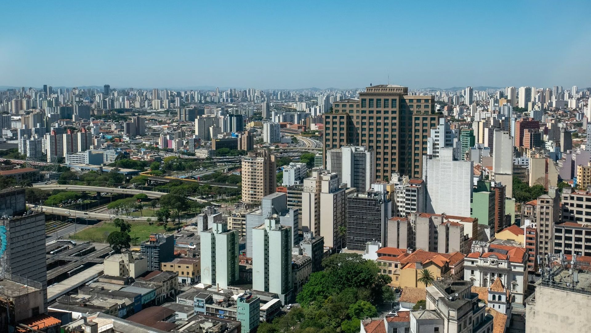 Prédio moderno branco de vários andares com fachada curva, varandas e lojas no térreo, situado em meio a árvores.