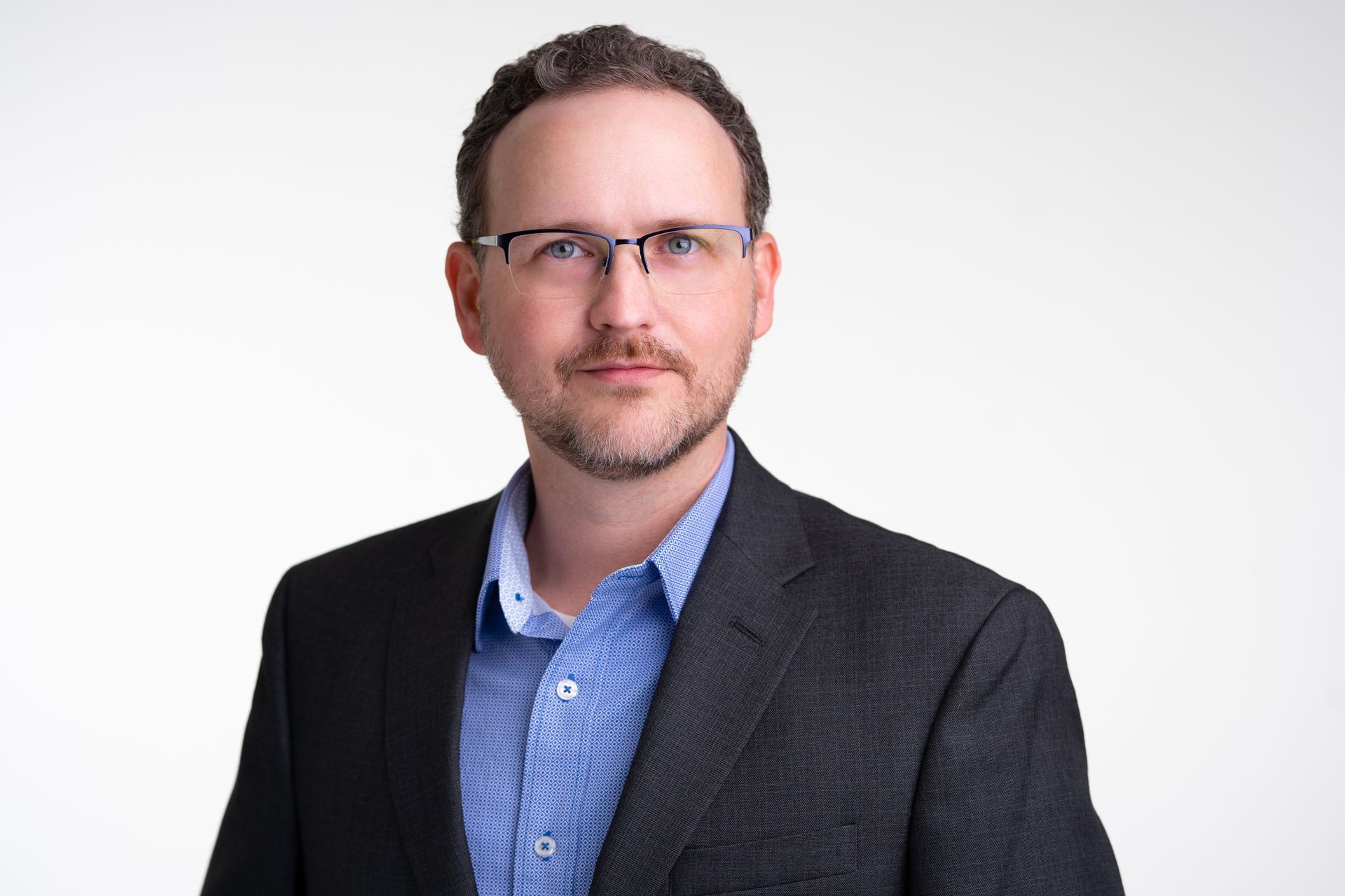 A man wearing glasses and a suit is standing in front of a white background.