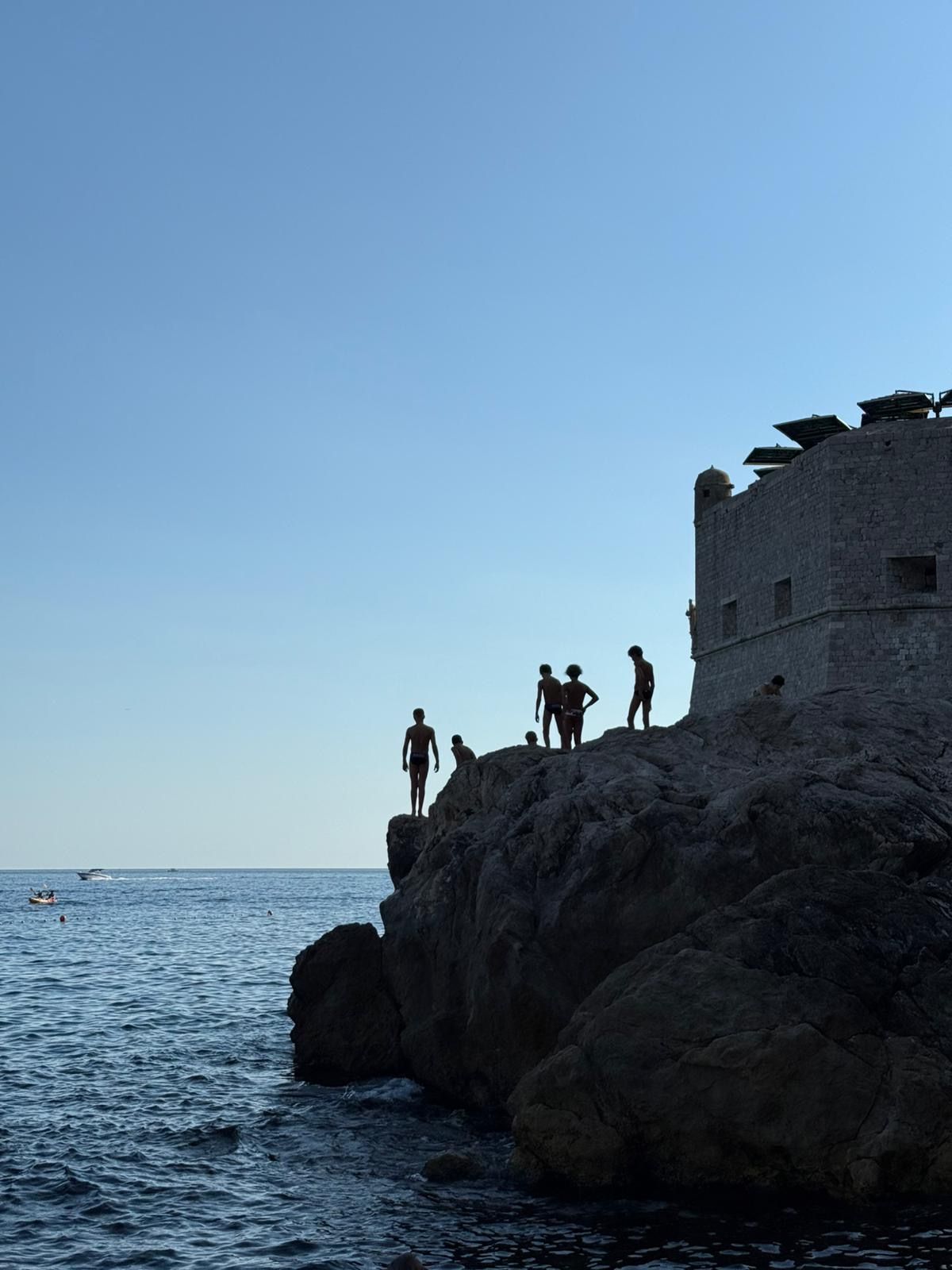 Silhouetted figures on a rocky cliff, preparing to jump into the sea, near a stone fortress.