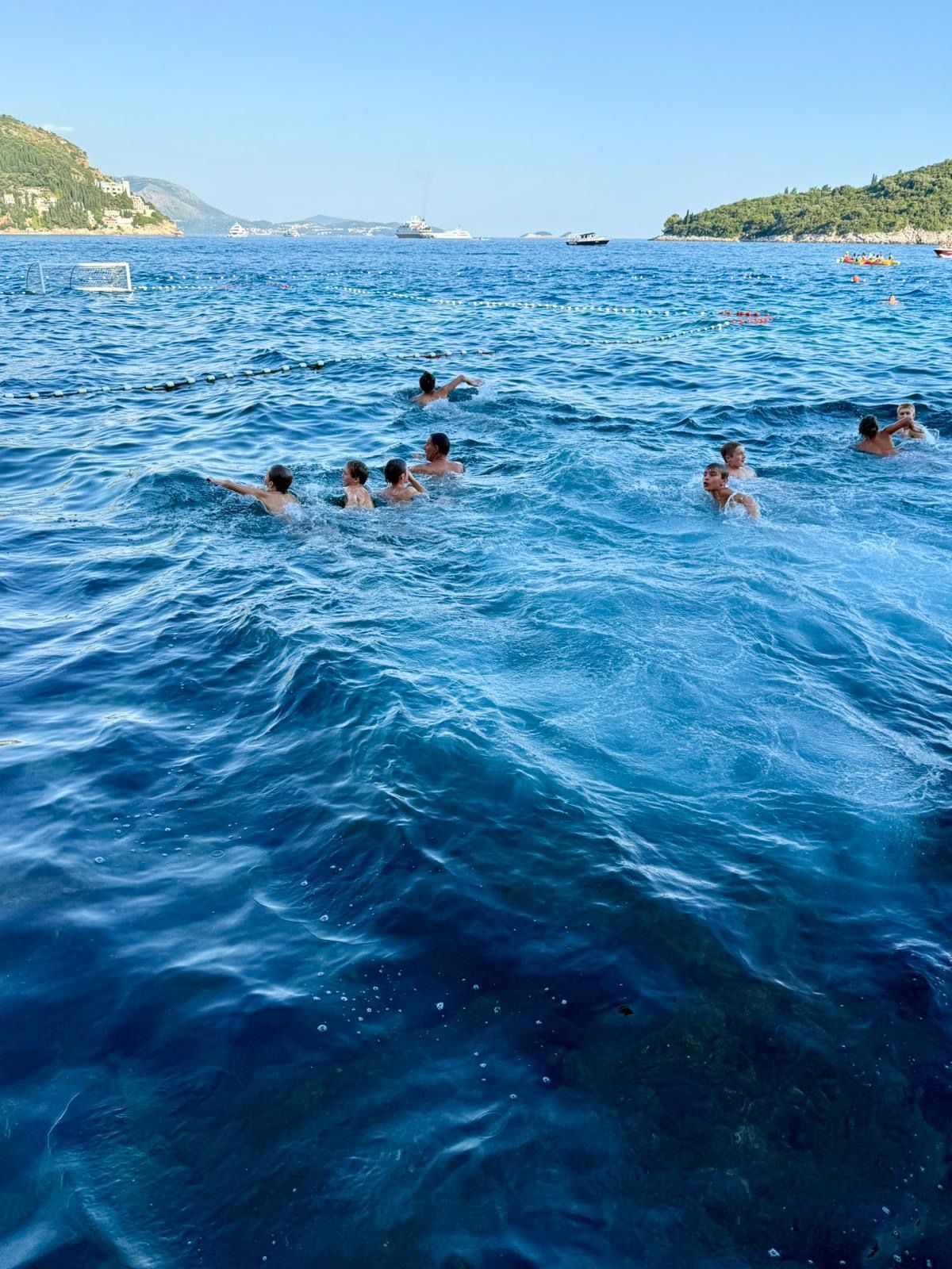 People swimming in the blue sea, with distant boats and shoreline under a clear sky.