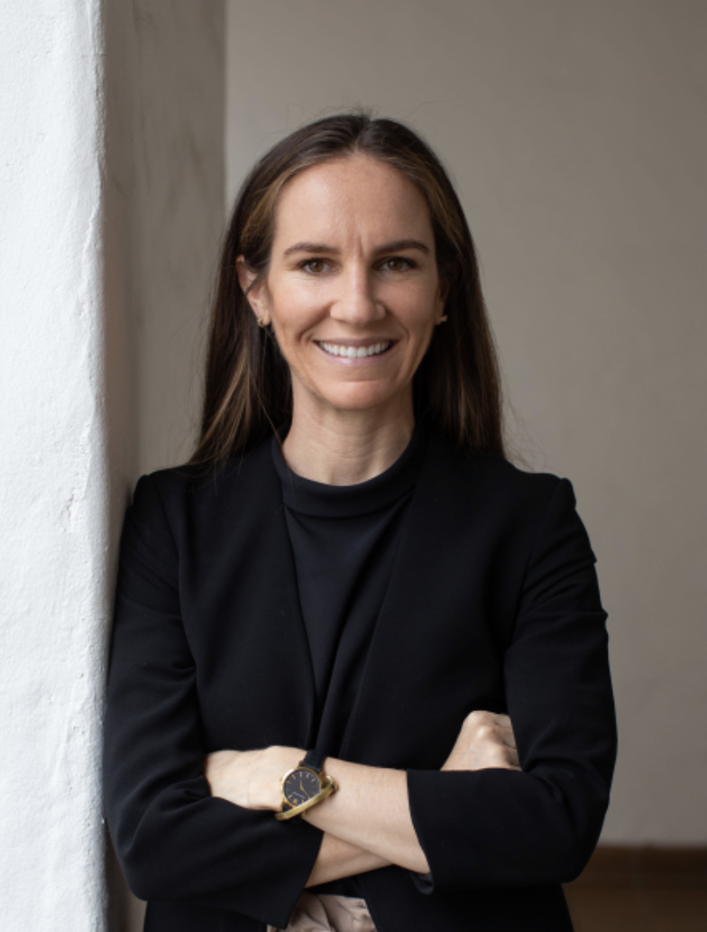A woman in a black jacket is leaning against a wall with her arms crossed and smiling.