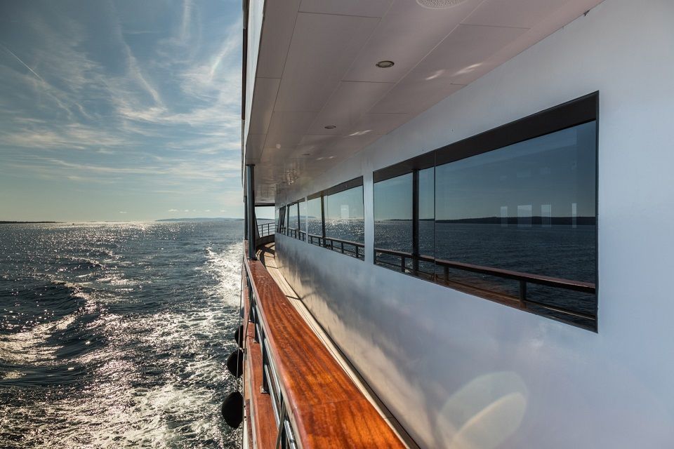 Side of a yacht with reflective windows sailing on a sunny day, water visible.