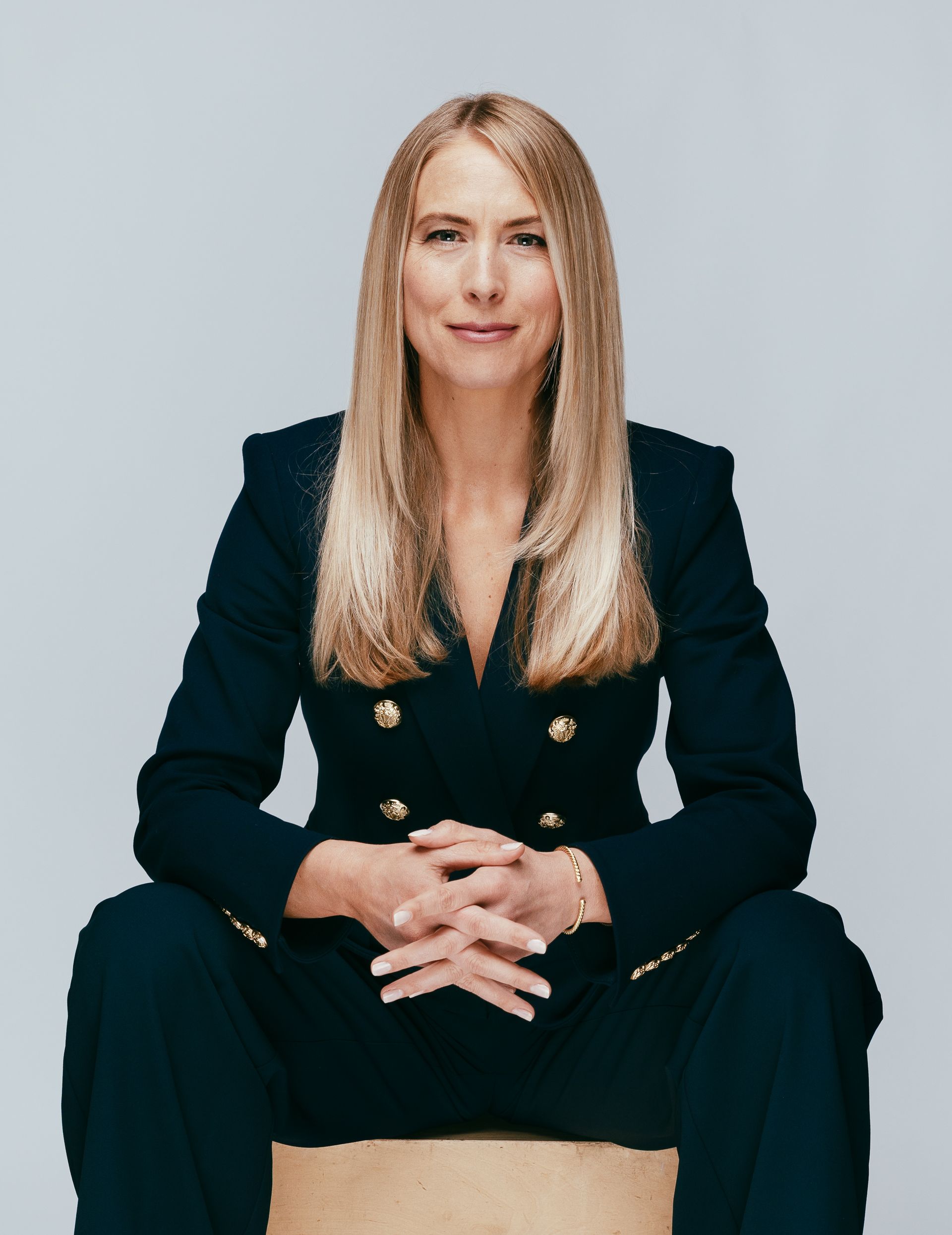 Woman with long blonde hair, wearing a navy suit, sits with hands clasped. Neutral background.