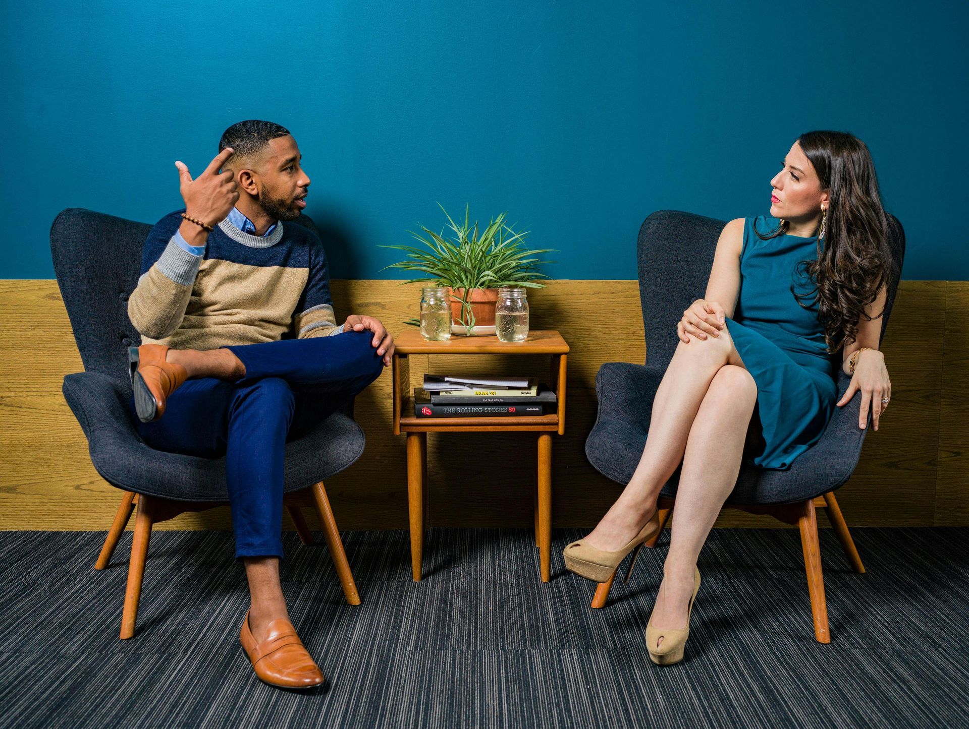 Man gesturing, woman listening in armchairs; small table with plant.