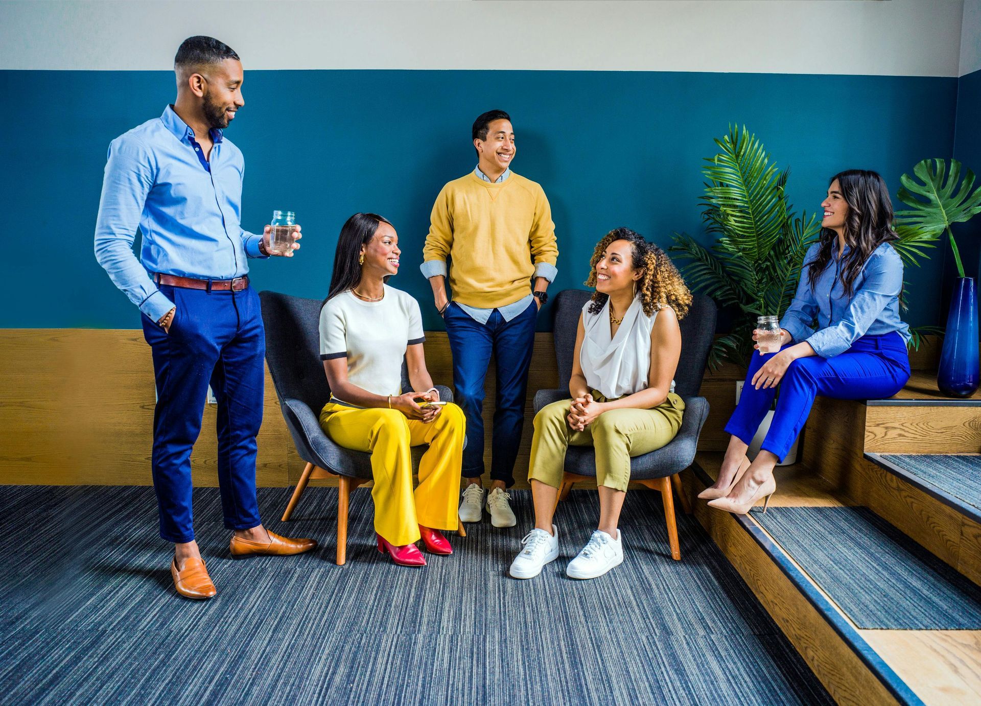 Group of diverse people socializing indoors, smiling, drinks in hand, in a modern setting with teal wall.