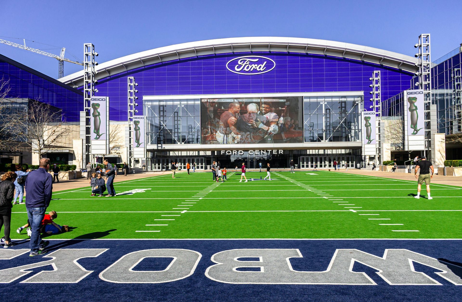 AT&T Stadium exterior with Ford logo, large screen, and field with Cowboys logo. People on the field.