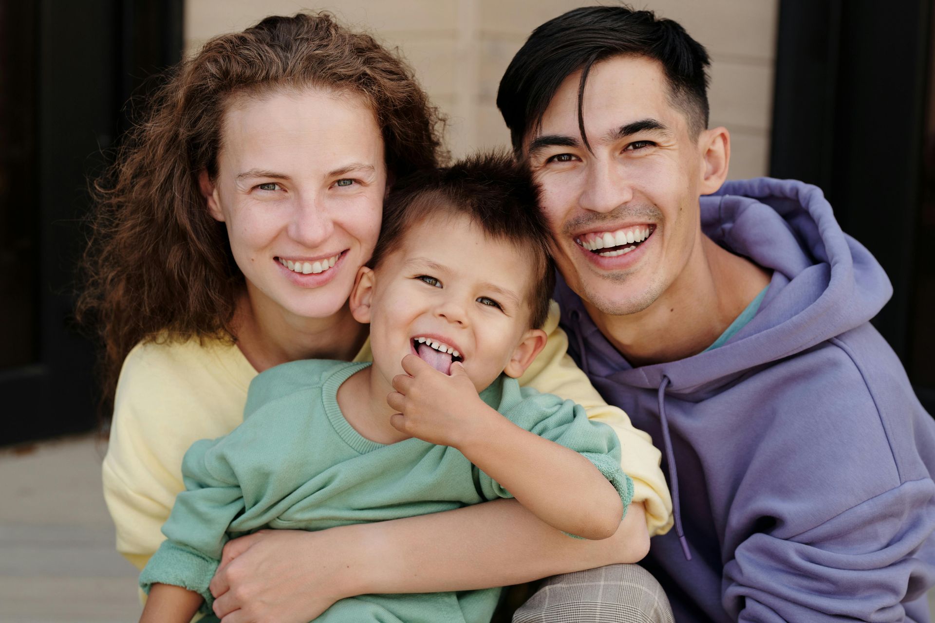 Family of three smiling, embracing outdoors. Woman in yellow, man in purple hoodie, child in green.
