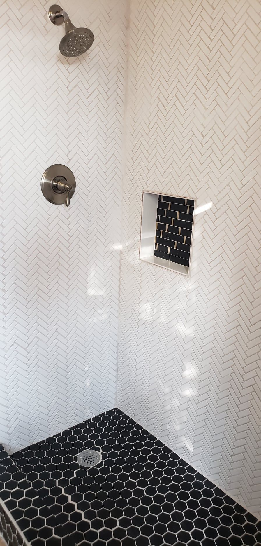 Empty shower stall with white textured walls, black hex tile floor, chrome fixtures, and a recessed shelf.