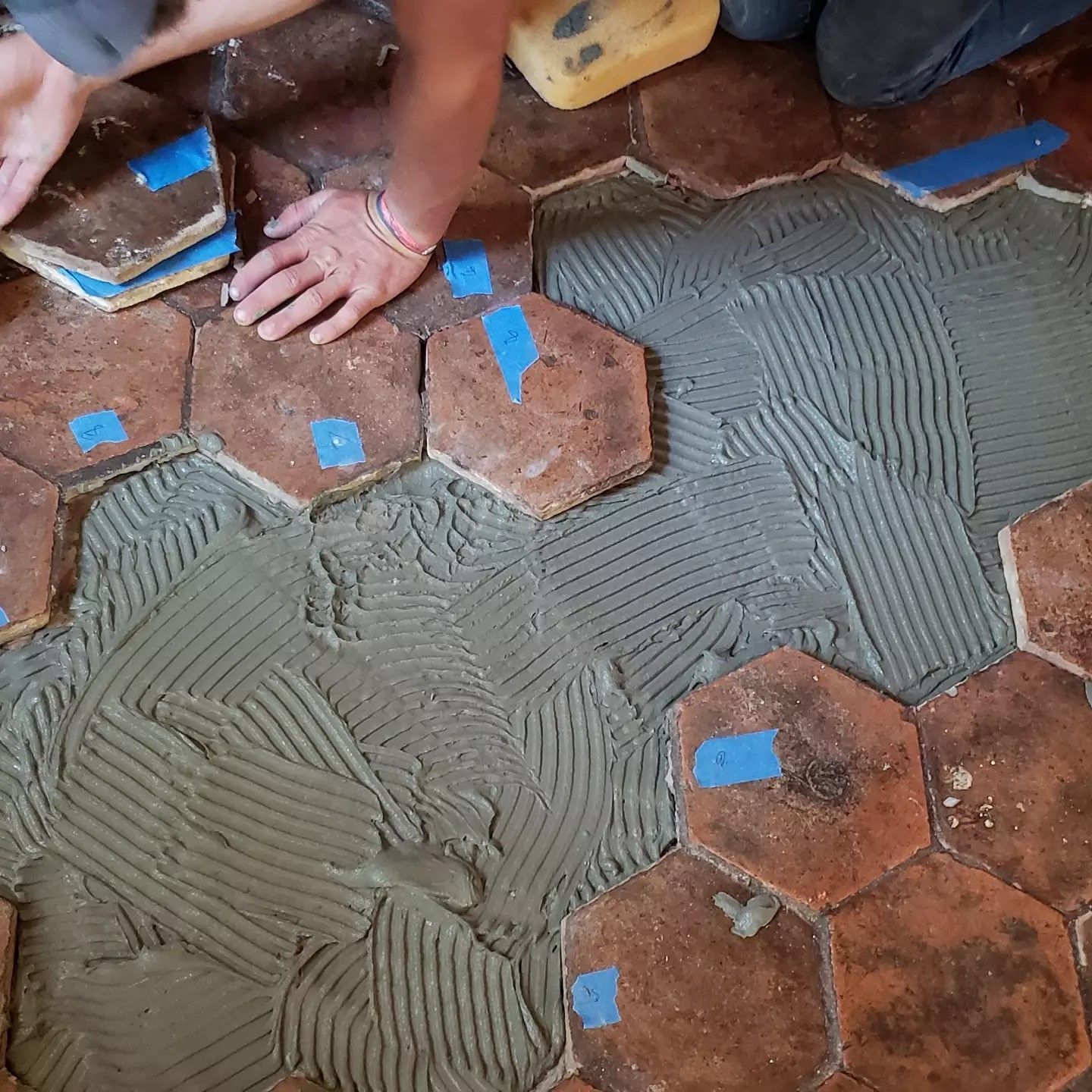 Hands spreading gray mortar over brown hexagonal floor tiles with blue spacers.