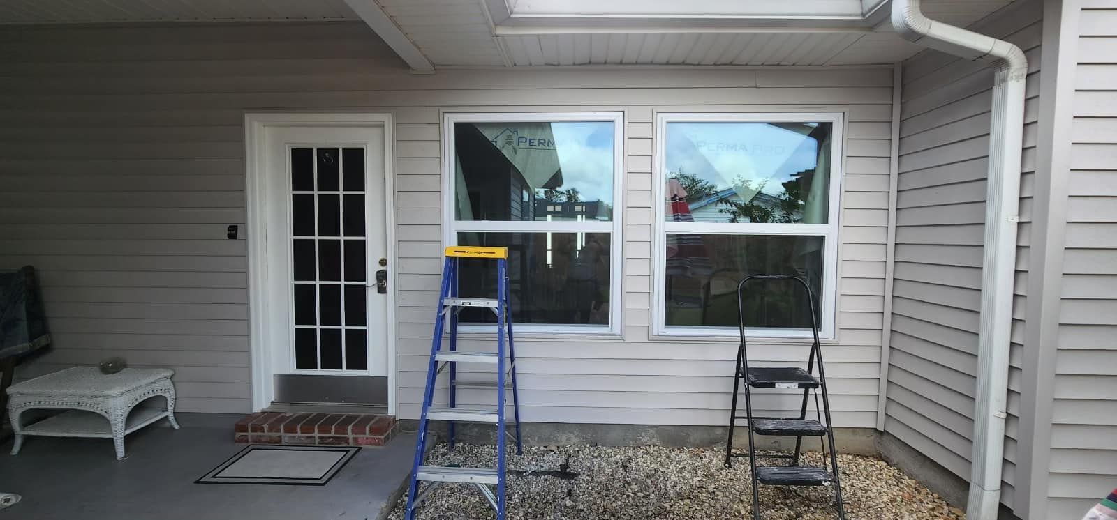 Home patio with a white door, large window, blue ladder, and tools against beige siding.
