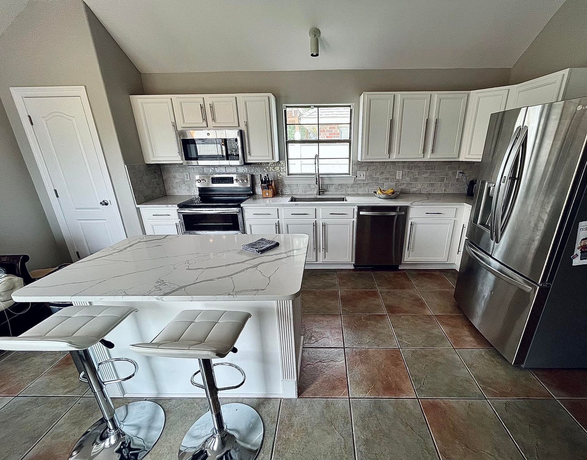 Bright kitchen with white cabinets, stainless fridge, island with stools, and tiled floor