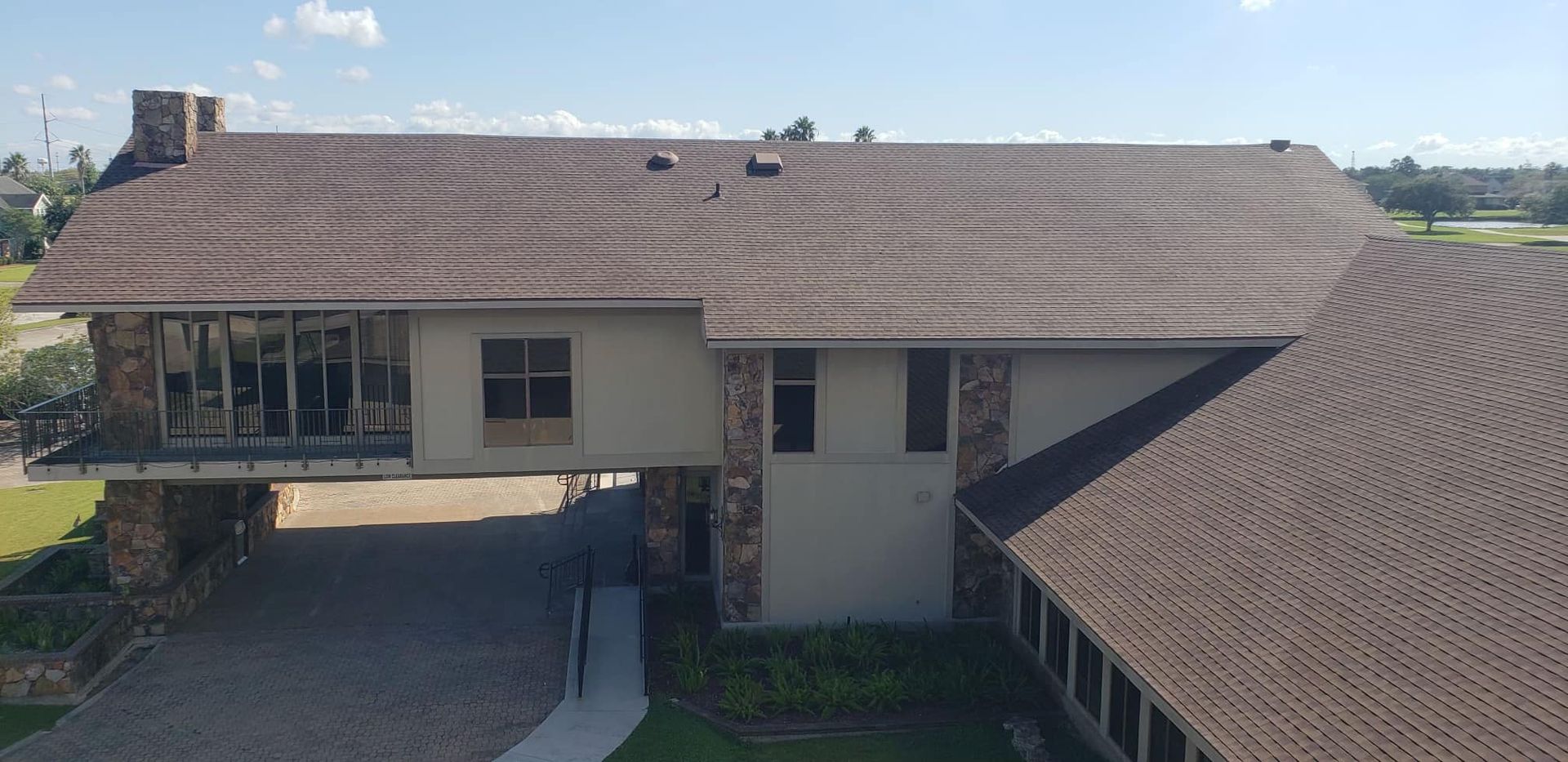 Aerial view of a beige house with a brown shingle roof and driveway, surrounded by open fields.