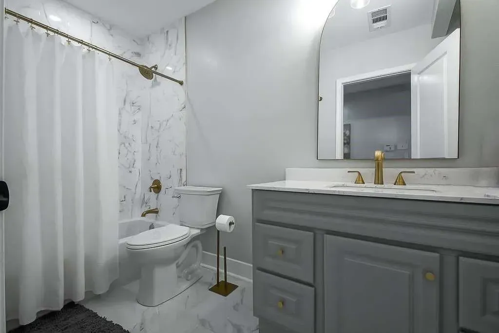 White bathroom with shower curtain, toilet, and gray vanity with mirror and gold faucet.