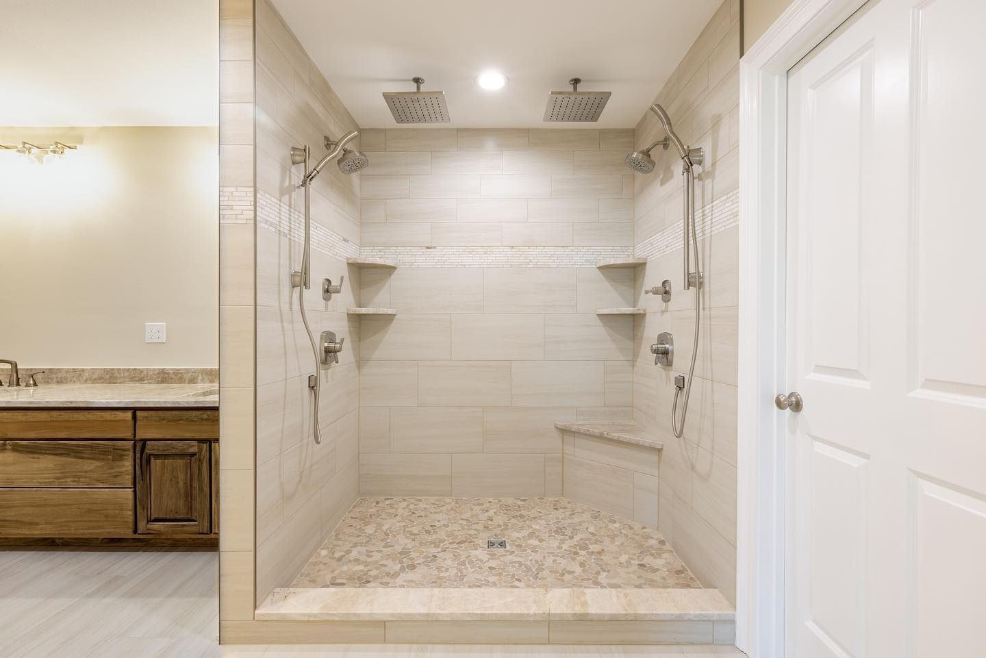 Walk-in shower with beige tile, built-in shelves, and pebble floor in a bright bathroom