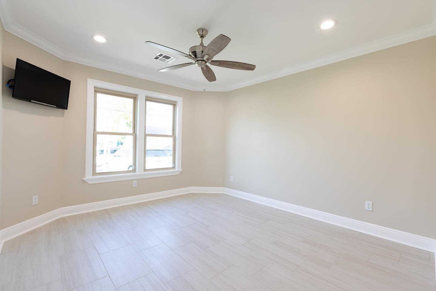 Bright empty bedroom with beige walls, light wood floor, ceiling fan, wall-mounted TV, and two windows
