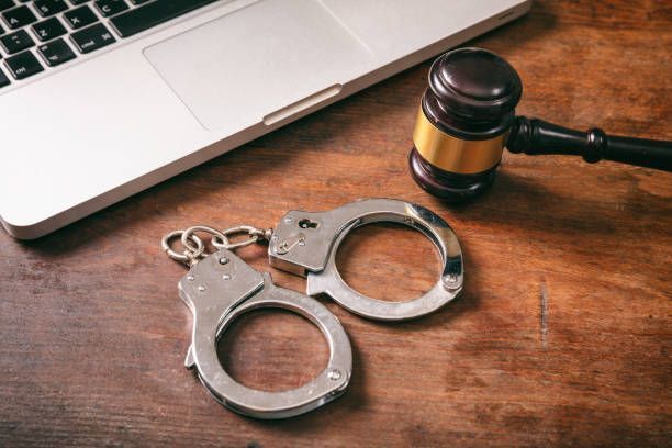 A pair of handcuffs and a judge 's gavel are on a wooden table next to a laptop.