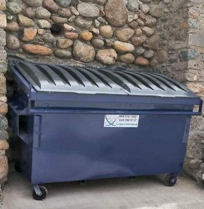 Contenedor de basura azul con tapa negra apoyado contra una pared de piedra.