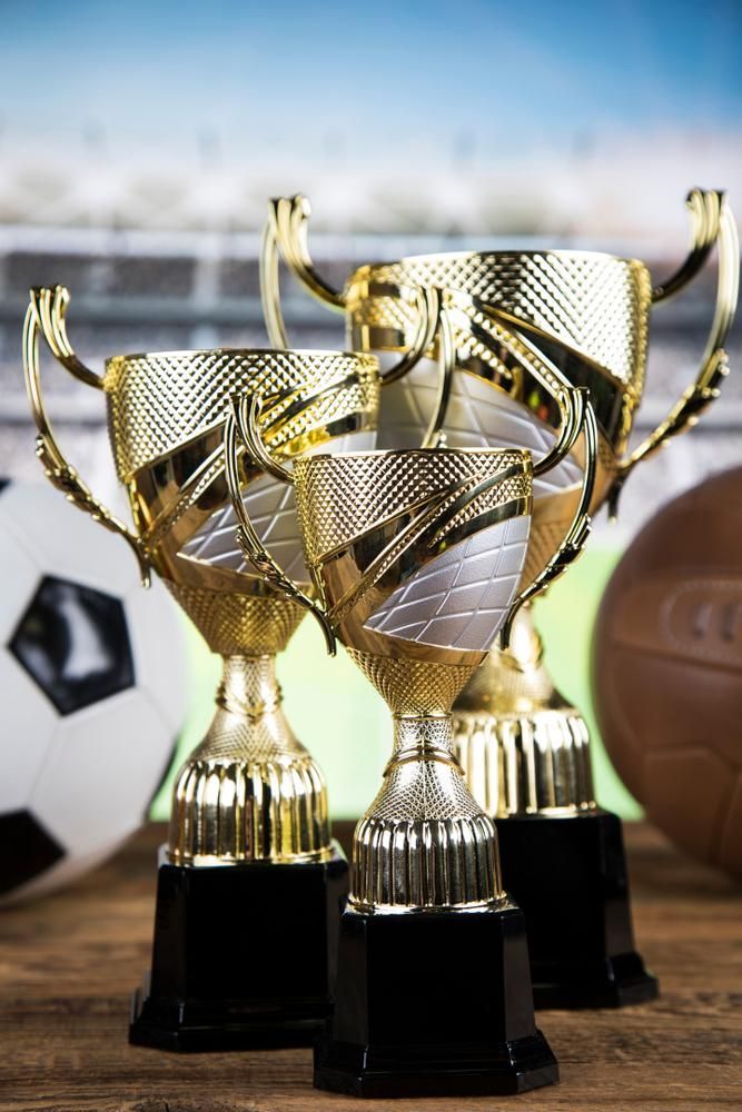 Three Gold Trophies Are Sitting on A Wooden Table Next to A Soccer Ball — Jimani Trophies & Engraving In Dapto, NSW