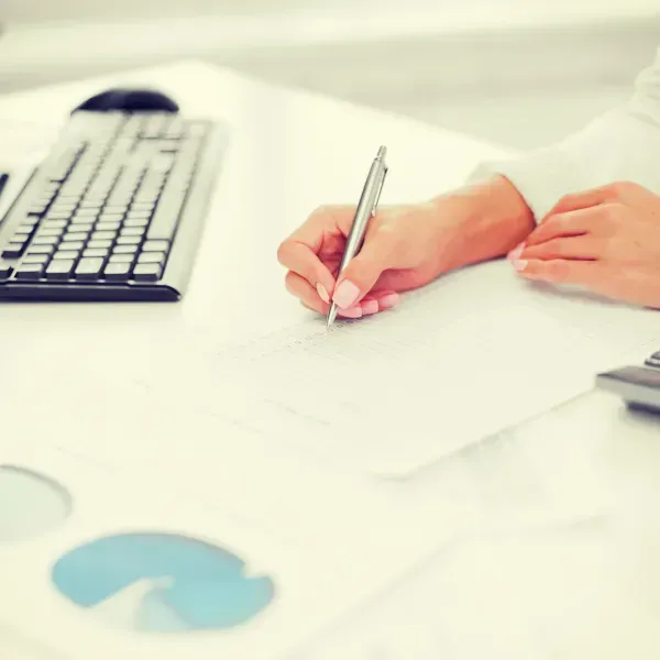 Person writing on paper with a pen at a desk, keyboard in view.