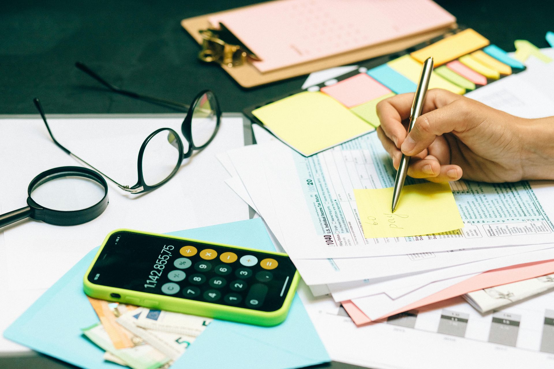 Hand writing on a form with pen, surrounded by papers, calculator, eyeglasses, and sticky notes.