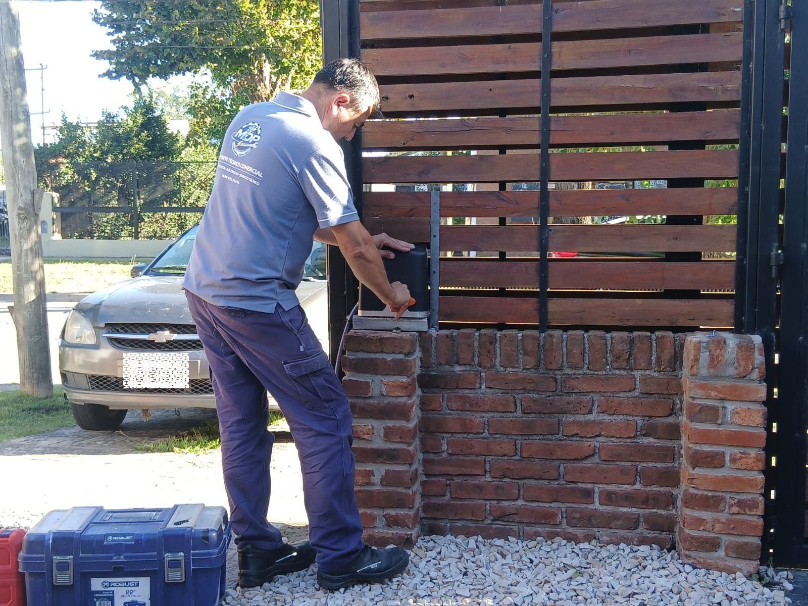Un trabajador con uniforme gris instala o repara un abrepuertas automático fijado a un pilar de ladrillo.