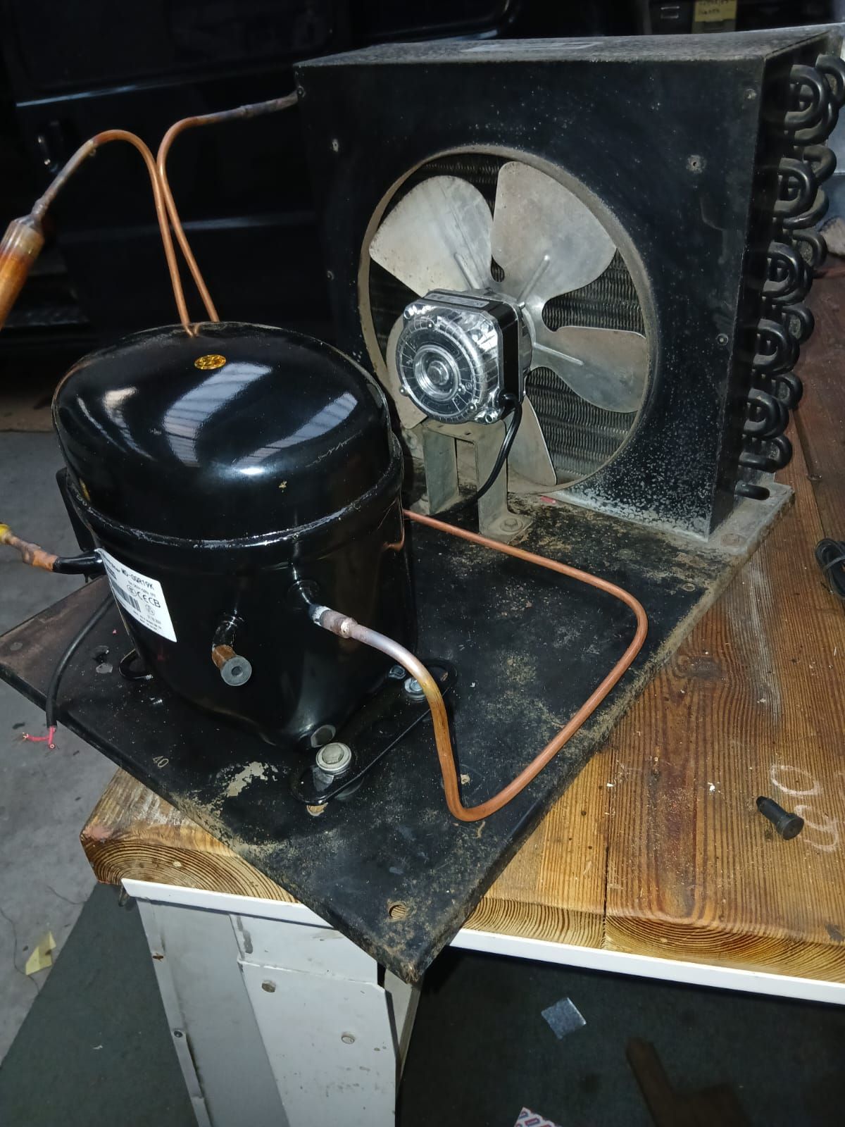 Un compresor de refrigeración negro conectado mediante tubos de cobre a una serpentina condensadora con un ventilador.