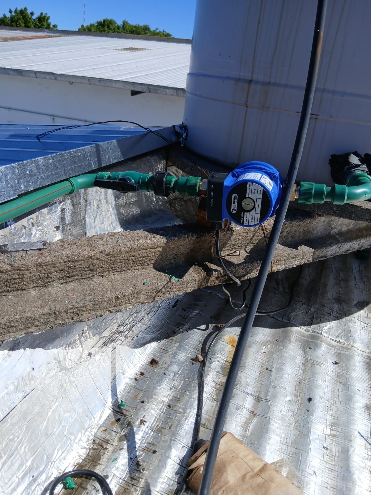 Una bomba de presión de agua azul conectada a tuberías verdes en una azotea junto a un gran tanque de agua de plástico.