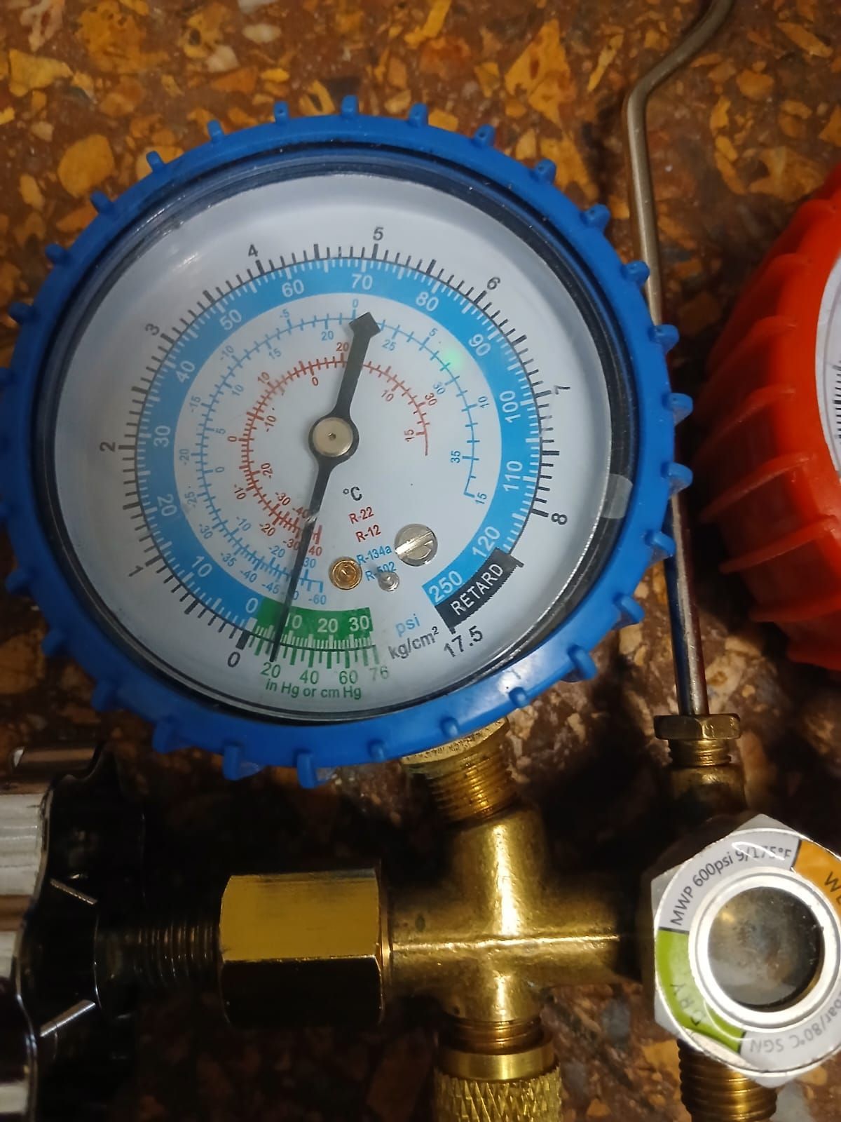 Un manómetro de refrigeración con carcasa azul y aguja que apunta a 75 PSI, montado sobre un colector de latón con mirilla.