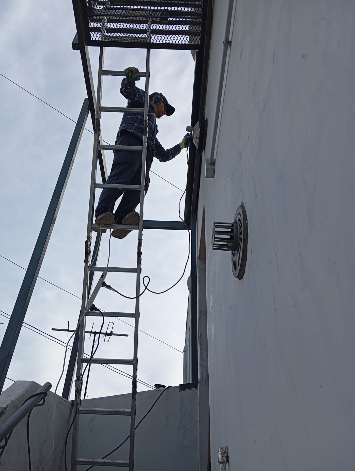 Un trabajador, subido a una escalera metálica alta, repara una luminaria fijada a una pared exterior blanca y alta.