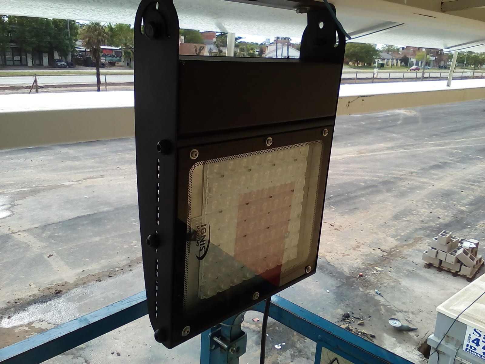 Un proyector LED industrial para exteriores montado sobre una estructura metálica, situado sobre una superficie de asfalto.