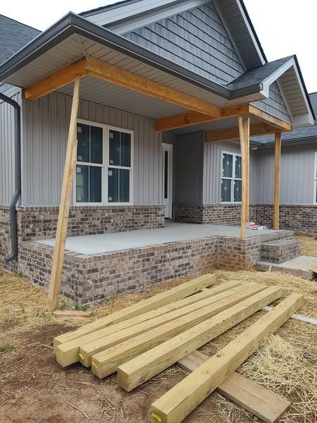 Front porch under construction; brick base, gray siding, wooden beams, and new lumber.