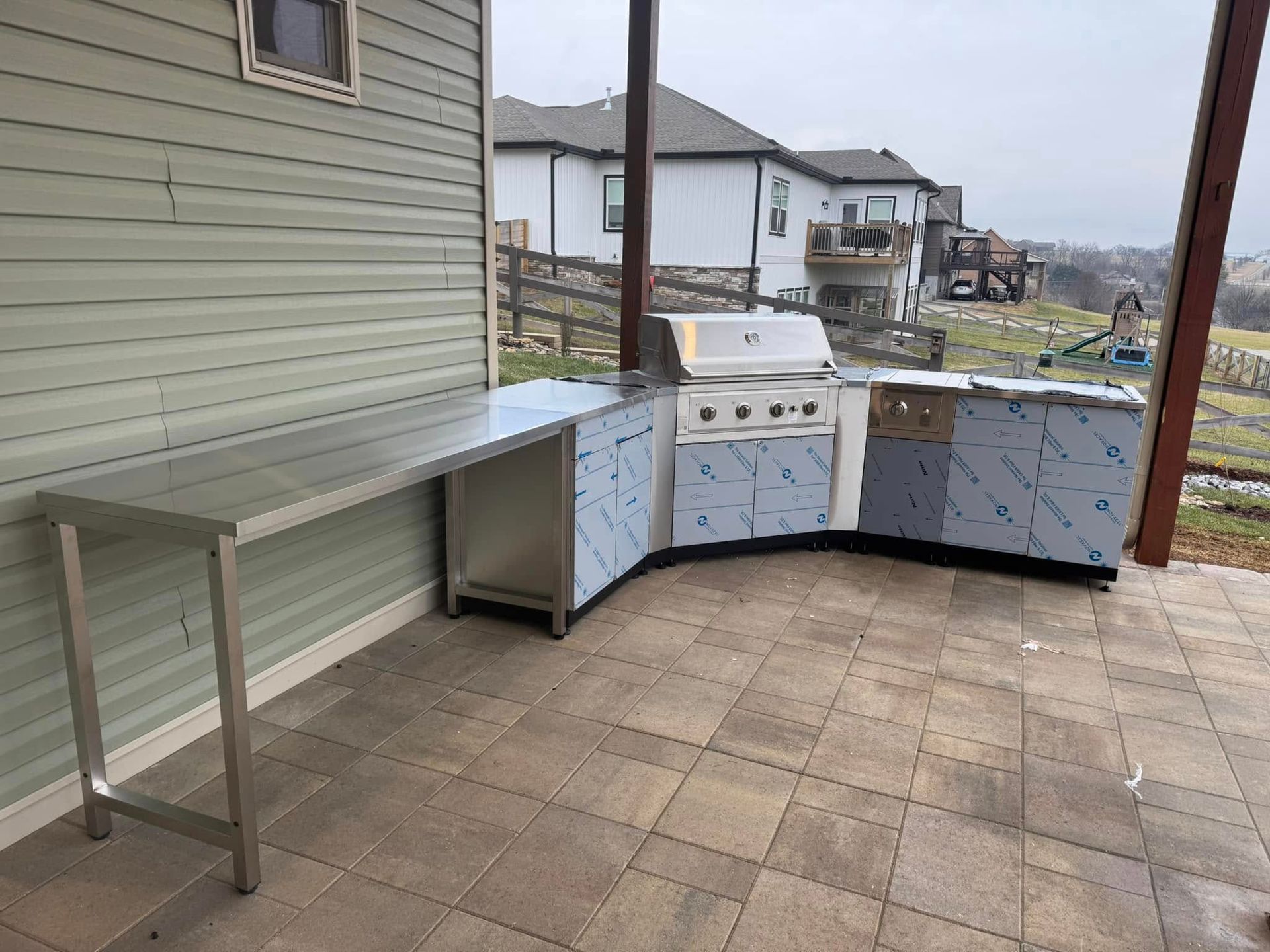 Outdoor stainless steel grill and counter on a patio, near a house and field, on a cloudy day.